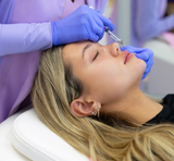 A close-up lifestyle shot of a woman receiving a brow tattoo or microblading treatment from a professional technician wearing purple scrubs and blue gloves.